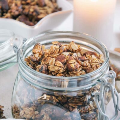 Lebkuchen-Granola in einem Einmachglas mit offenem Deckel. Dekoriert mit einer Kerze und mehr Granola in einer kleinen Sternenschale im Hintergrund. Aufnahme im 45-Grad-Winkel.