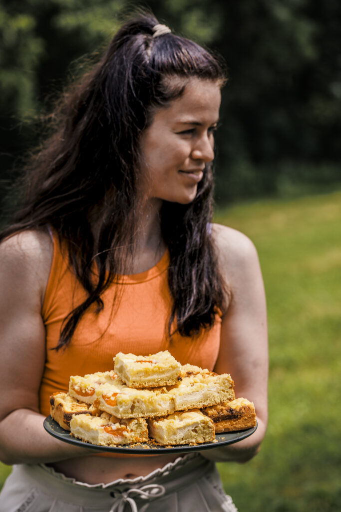 Foobloggerin Sarah von Oats and Crumbs hält einen Teller mit einigen Sücken Marillen-Topfenkuchen mit Streuseln.